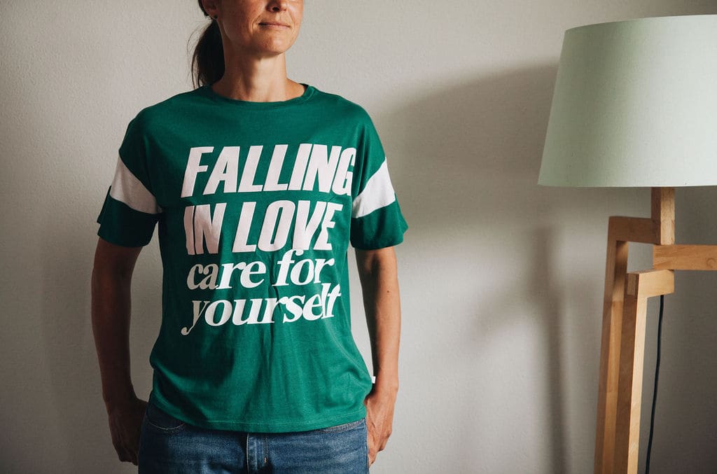 Oberkörper einer Frau, die ein T-Shirt trägt auf dem steht: Falling in love care for yourself.