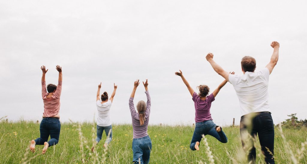 nach dem Coaching nach The Work of Byron Katie springen fünf Menschen, die von hinten sichtbar sind auf einer Wiese in die Höhe und heben die Arme glücklich und befreit nach oben