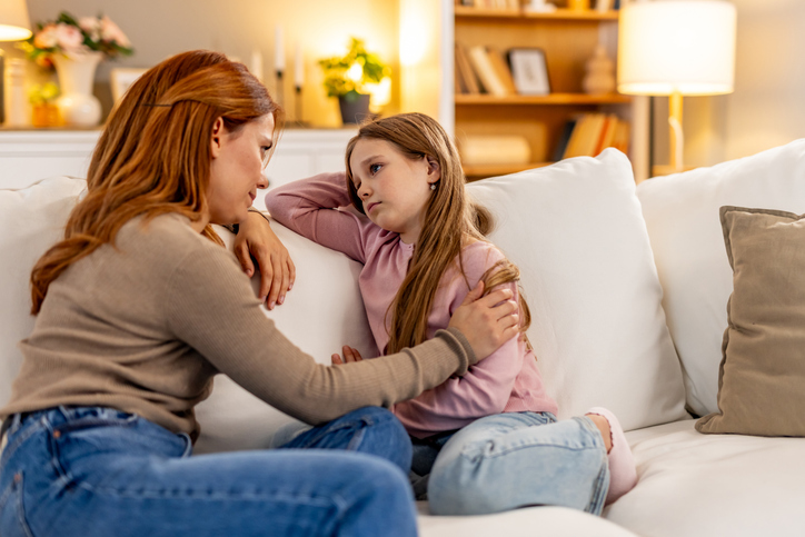 Eine Mutter gibt ihrer Tochter Liebe und Sicherheit zuhause auf einer weißen Couch im gemütlichen Wohnzimmer